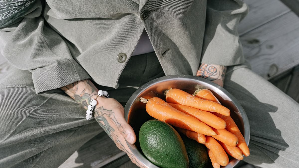 Person in gray suit holding a bowl of fresh carrots and avocados outdoors.